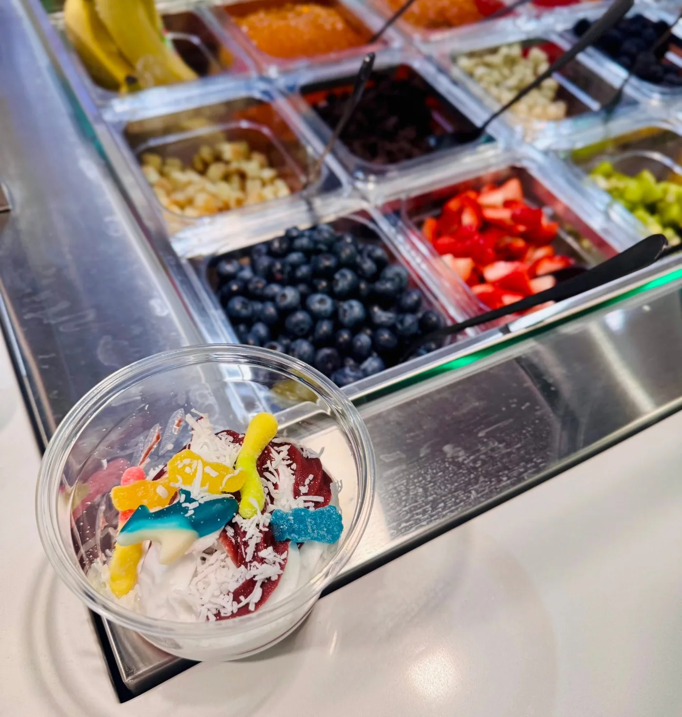 Frozen yogurt cup with gummy sharks, gummy worms and coconut beside the toppings bar